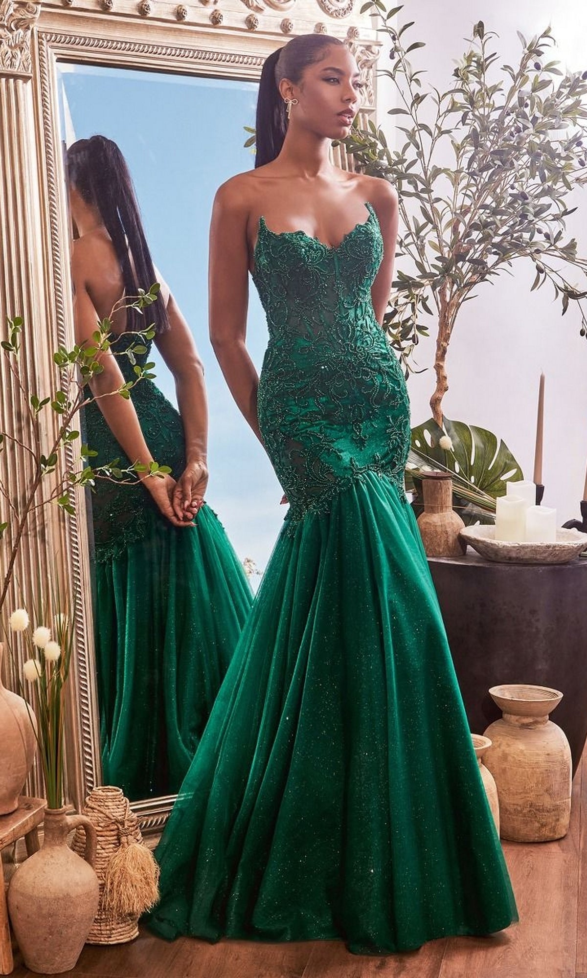 A woman in the Strapless Long Lace Mermaid Prom Dress CDS482, an emerald green fitted gown, stands before a large mirror amid potted plants and decorative vases.